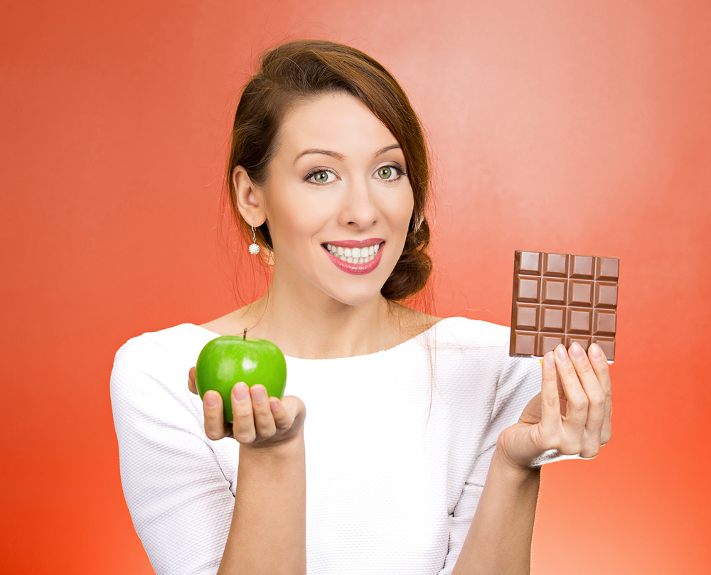 Closeup portrait, beautiful, smiling young woman offering nutritious lime apple as an alternative to unhealthy square milk chocolate, isolated red background. Food diet option situations, dilemma.