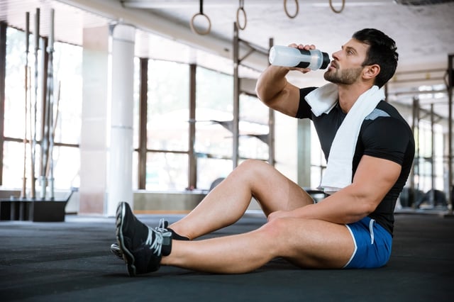 Fitness man wearing blue shorts and black t-shirt sitting on the floor and drinking water.jpeg